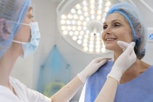 médico passa a mão sobre o rosto de um mulher já preparada para a cirurgia facial